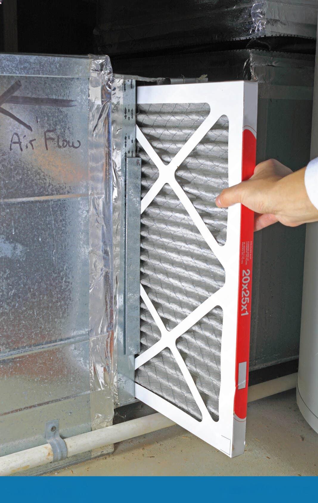 Close-up of a person’s hand sliding a 20x25x1 pleated air filter into a metal HVAC return duct, with an airflow arrow marked on the ductwork.